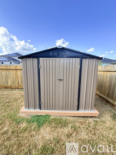 A small, grey and blue shed sits on a wooden platform in a grassy backyard.