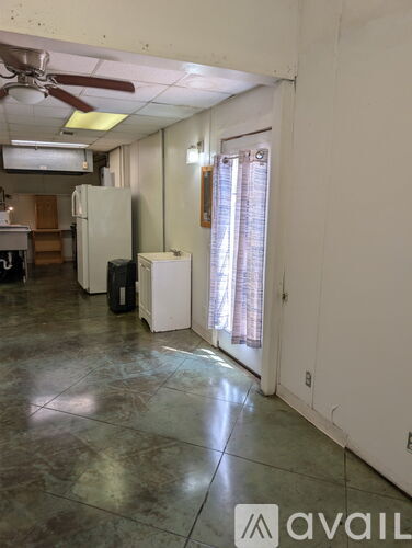 A room with a fan, a refrigerator, and a window with curtains.