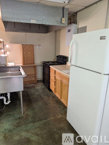 A white refrigerator stands in a kitchen with wooden cabinets and a stainless steel sink.