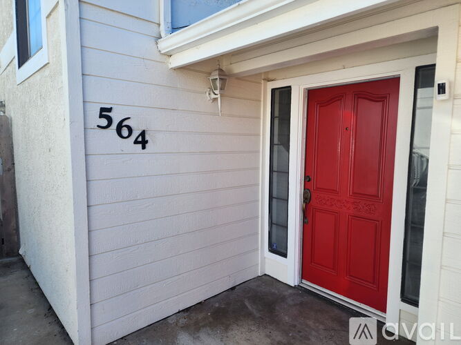 A red door is on the right side of a house with the number 564 on the wall.