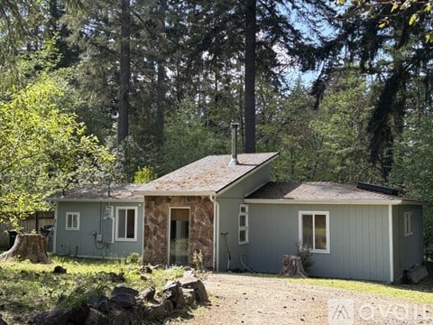 A small house with a grey exterior is surrounded by trees and rocks.