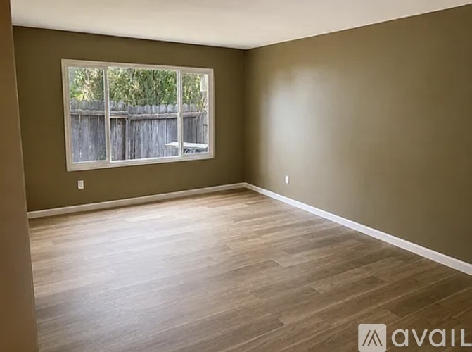 A room with brown walls and wooden flooring, with a window showing a wooden fence and greenery outside.