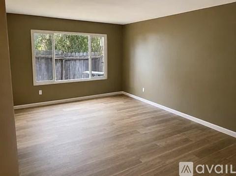 A room with brown walls and wooden flooring, with a window showing a wooden fence and greenery outside.