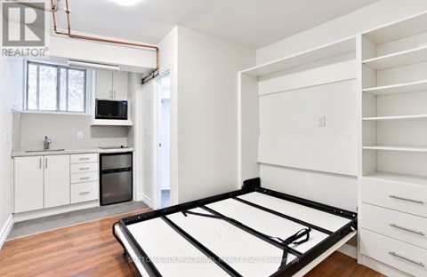 A bed frame is in the middle of a white room with a kitchen in the background.