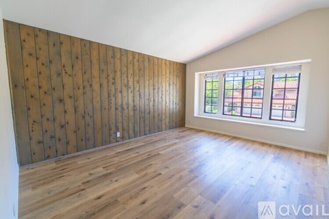 A room with wooden flooring and a wooden wall panel with a window showing a view of a building.