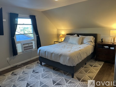 A bedroom with a large bed, a window with blinds, and a nightstand with a lamp.