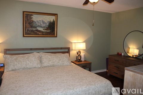 A bedroom with a bed, a painting on the wall, a side table with a lamp, and a dresser with a mirror.