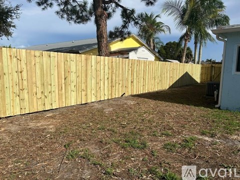 A wooden fence in a yard with a house and palm trees in the background.
