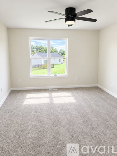 A room with a carpet floor, a ceiling fan, and a window showing a view of a house and trees outside.