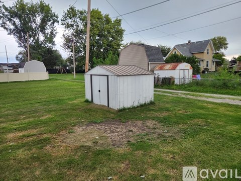 A shed sits in a grassy field with a house and trees in the background.
