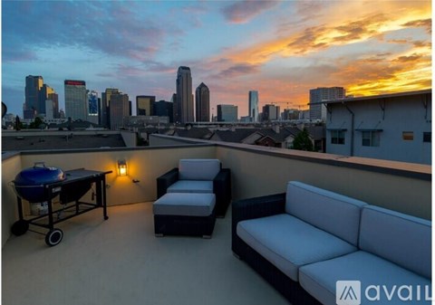 A balcony with a table, chairs, and a sofa overlooking a city skyline at sunset.