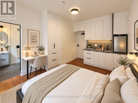 A bedroom with a bed, desk, and kitchen area.