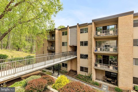 A bridge connects two buildings in a courtyard with bushes and trees.