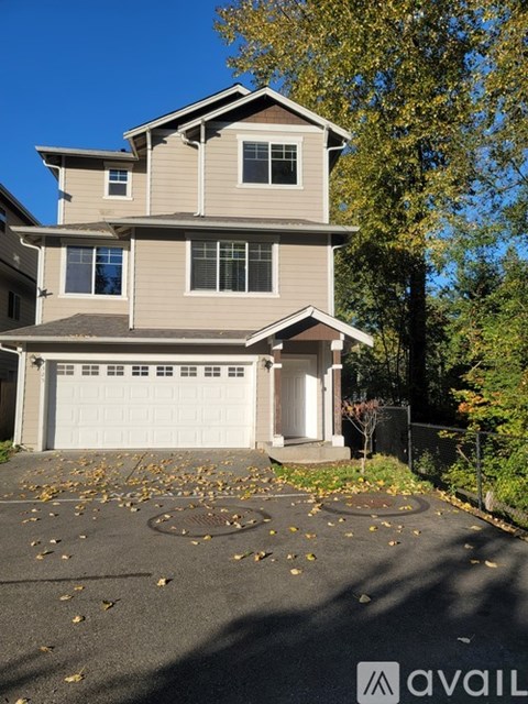 A two-story house with a garage is surrounded by trees and has fallen leaves on the driveway.