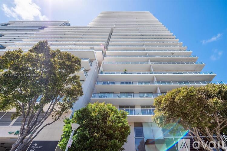 A tall white building with balconies on each floor is surrounded by trees.