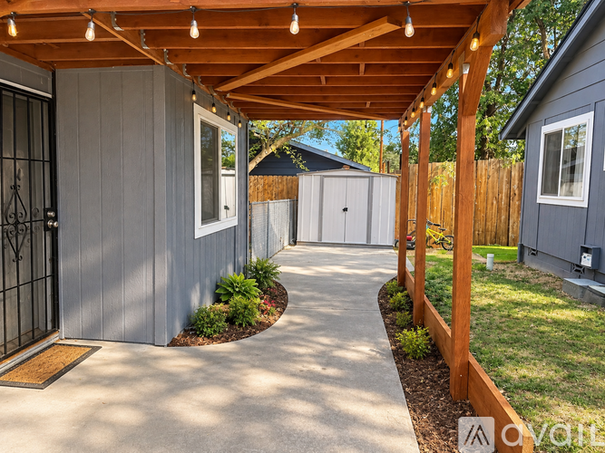 A wooden pergola with string lights is over a concrete walkway leading to a grey house.