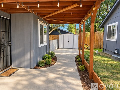 A wooden pergola with string lights is over a concrete walkway leading to a grey house.