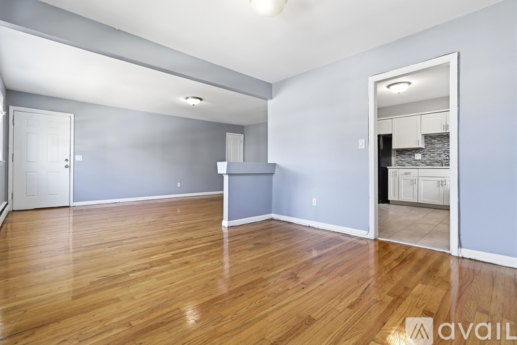 A spacious room with wooden flooring and a kitchen visible through the open door.