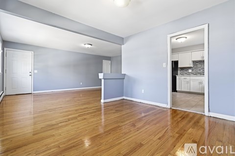 A spacious room with wooden flooring and a kitchen visible through the open door.