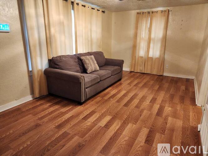 A brown couch sits in a room with wooden flooring and curtains.