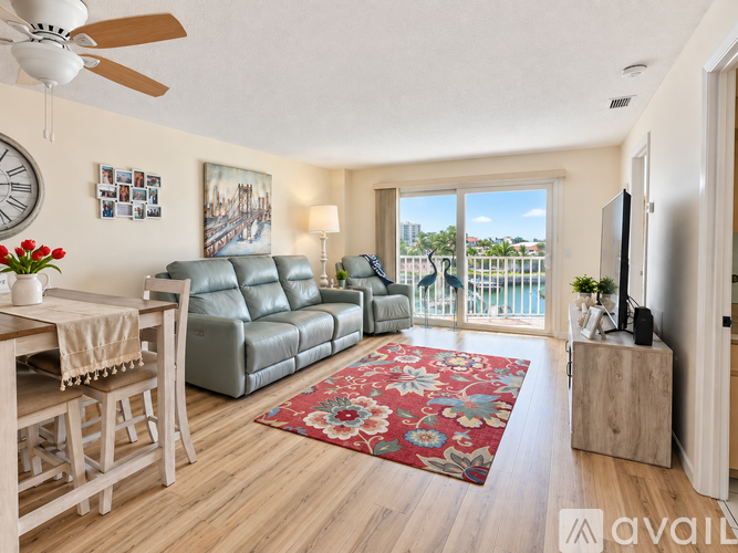 A living room with a grey leather sofa, a wooden dining table with chairs, a red floral rug, a ceiling fan, and a view of a balcony with a railing.