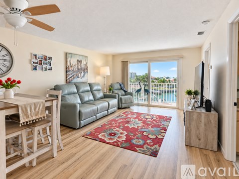 A living room with a grey leather sofa, a wooden dining table with chairs, a red floral rug, a ceiling fan, and a view of a balcony with a railing.