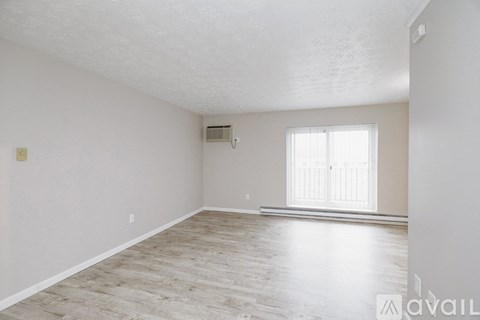 A spacious room with light wood flooring and a window with blinds.