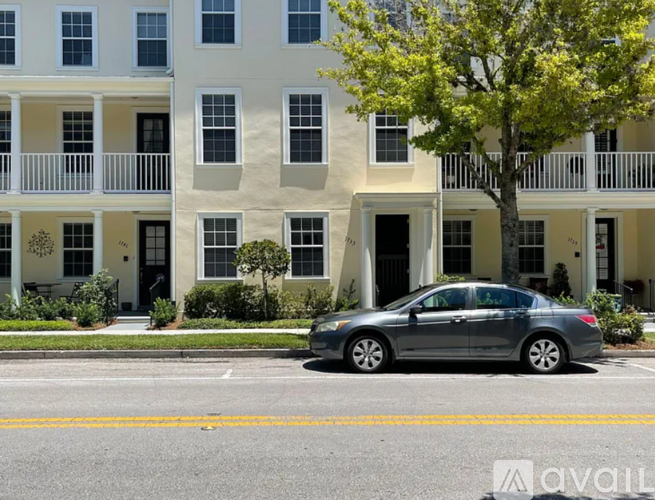 A silver car is parked in front of a white building with a tree in front of it.