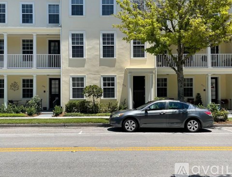 A silver car is parked in front of a white building with a tree in front of it.