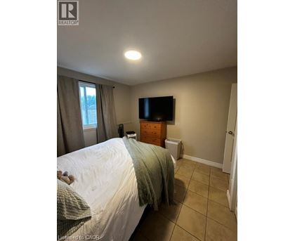 A bedroom with a bed, a window with curtains, a dresser with a flat screen TV mounted above it, and a white wall.