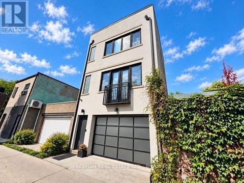 A two-story building with a garage door and a balcony is surrounded by greenery.