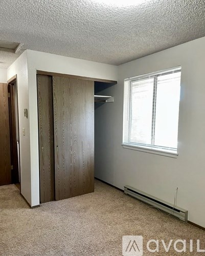 A room with a carpeted floor, a window with blinds, and a wooden wardrobe.