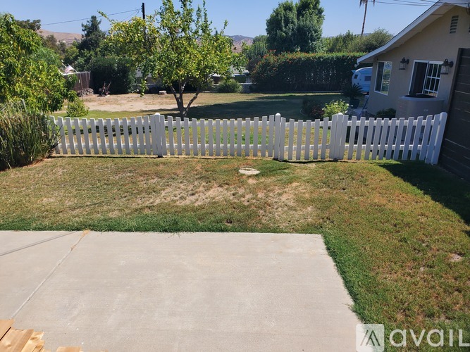A white picket fence encloses a yard with a tree and a house in the background.