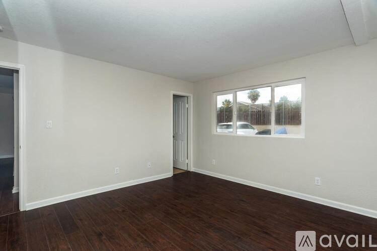 A room with wooden floors and white walls, with a window showing a view of a fence and trees.