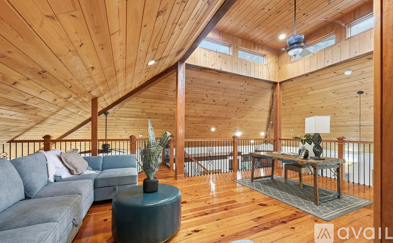 A living room with wooden walls and ceiling, a grey sofa, a round table with a plant on it, and a balcony with a railing.