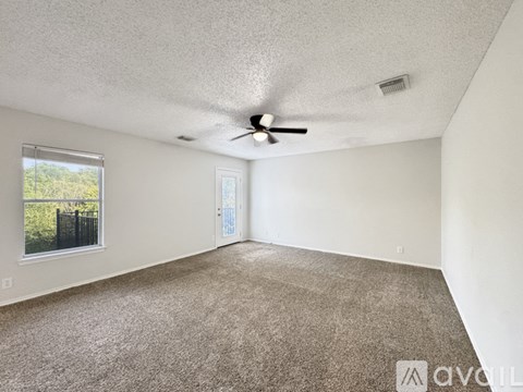 A room with a carpeted floor, a ceiling fan, and a window with a view of trees and a fence.