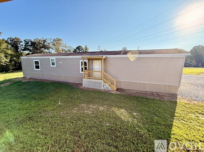 A mobile home with a brown siding and a white door is surrounded by a gravel driveway and a grassy lawn.