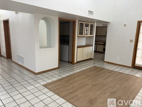 A room with a wooden floor and white walls, with a kitchen area visible through an open door.