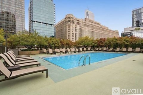 A pool surrounded by lounge chairs on a patio with trees and buildings in the background.