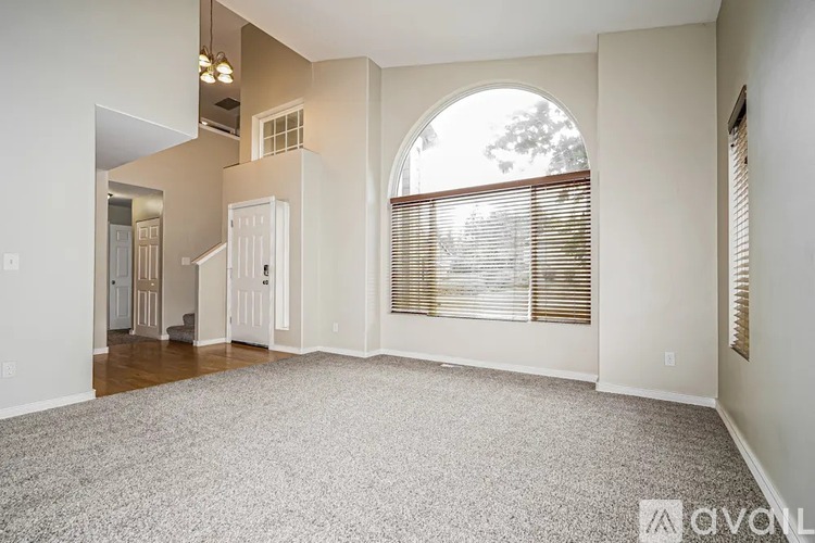 A spacious living room with a carpeted floor and a large arched window with blinds.