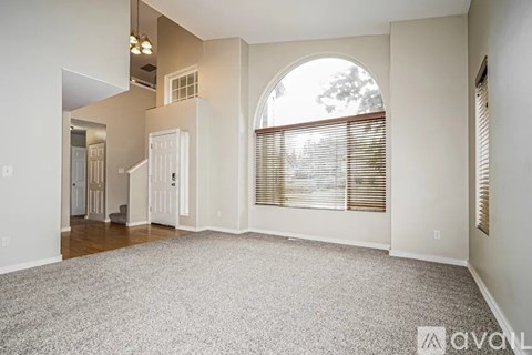 A spacious living room with a carpeted floor and a large arched window with blinds.