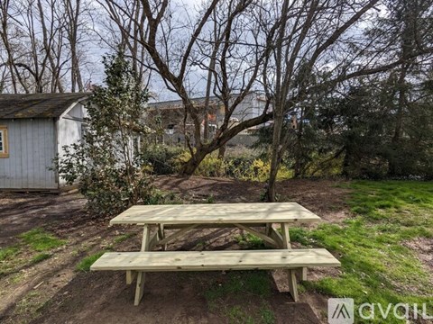 A wooden picnic table is in the foreground of a grassy area with a house and trees in the background.