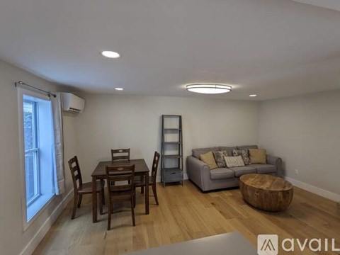 A living room with a grey couch, a wooden coffee table, a dining table with chairs, and a bookshelf.