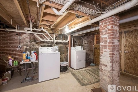 A kitchen area with a white fridge and dishwasher in a room with exposed pipes and brick walls.