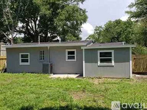 A grey house with a white door and windows is surrounded by green grass and trees.