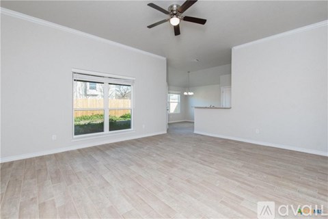 A spacious room with a ceiling fan and light, hardwood flooring, and a large window with a view of a fence and greenery outside.