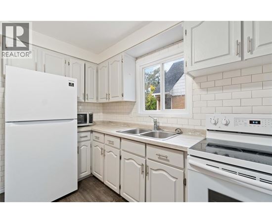 A white refrigerator stands in a kitchen with white cabinets and a white stove.