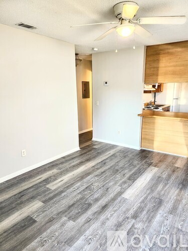 A room with a ceiling fan and a wooden floor leading to a kitchen.