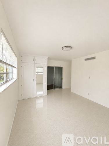 A spacious, empty room with white walls and flooring, a window with blinds, and a white cabinet with glass doors.