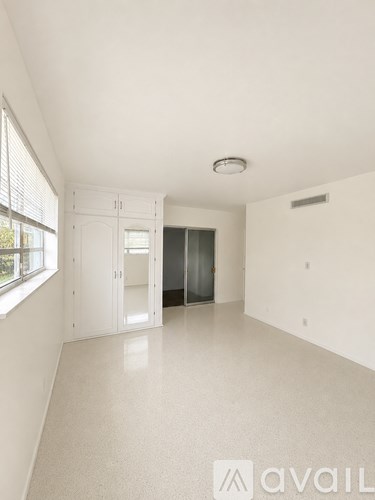 A spacious, empty room with white walls and flooring, a window with blinds, and a white cabinet with glass doors.